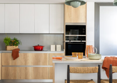 Cocina abierta con isla central en madera y blanco, lámparas colgantes verdes y taburetes de enea.