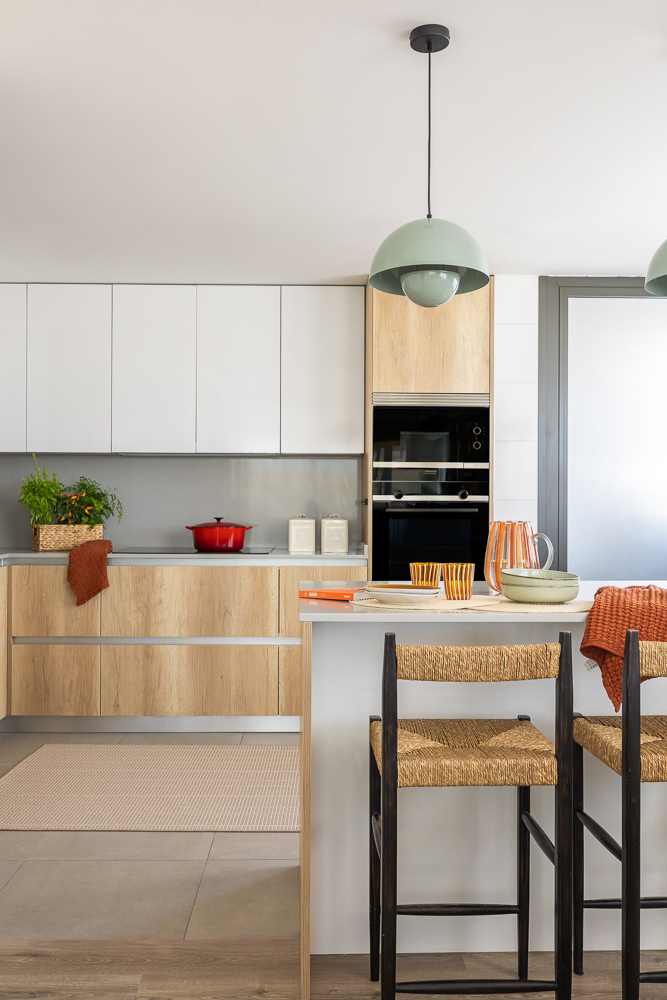 Cocina abierta con isla central en madera y blanco, lámparas colgantes verdes y taburetes de enea.