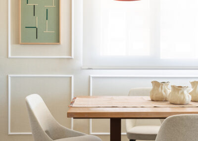 Comedor con mesa de madera, sillas tapizadas en beige, lámpara colgante roja y obra de arte geométrica en la pared.
