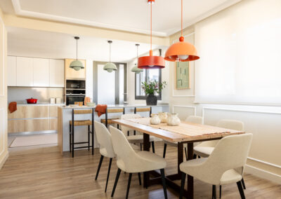 Comedor con mesa de madera y lámparas colgantes de colores, integrado con la cocina abierta en madera y blanco.