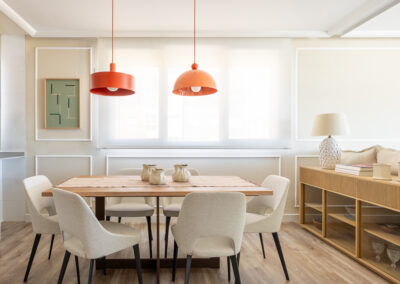 Comedor con mesa rectangular de madera, sillas tapizadas en beige y dos lámparas rojas colgantes.