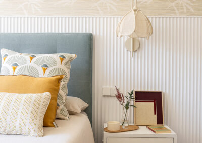 Dormitorio diseñado por María del Valle Interiorismo con cabecero azul, cojines decorativos y papel pintado de hojas en tonos neutros.
