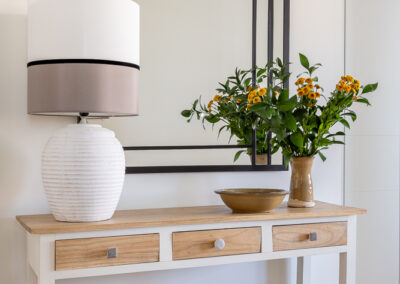 Hall de vivienda con consola de madera y blanco, espejo con marco geométrico y jarrón con flores.