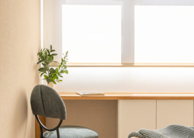 Rincón de teletrabajo con escritorio a medida en madera, silla tapizada en verde y ventana con estor blanco.