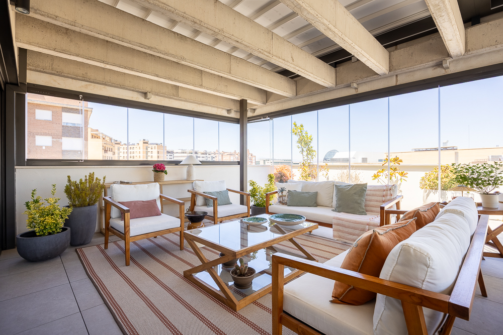 María_del_Valle_Interiorismo_terraza_ático_vistas_sofás_cojines_alfombra_exterior Terraza acristalada con sofás y cojines decorativos en tonos cálidos y verdes.