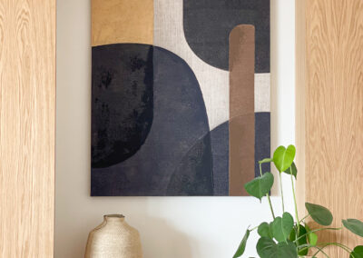 Cuadro abstracto en tonos tierra con planta decorativa junto a mueble de madera.