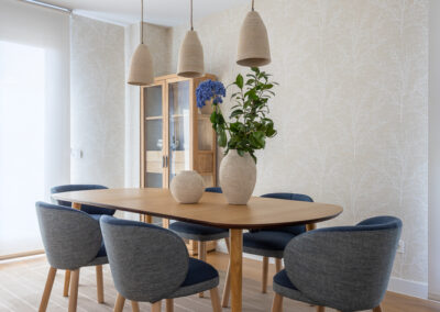 Comedor con mesa de madera, sillas tapizadas en tonos grises y azules, lámparas colgantes y papel pintado en tonos neutros.