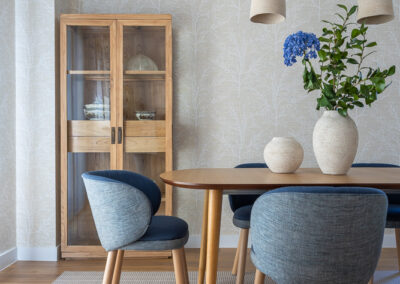 Comedor con mesa de madera, sillas tapizadas en azul, vitrina de madera y papel pintado en tonos neutros con textura vegetal.