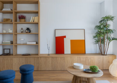 Salón con mueble bajo de madera a medida, estantería abierta, cuadro apoyado en tonos rojos y naranjas y decoración minimalista.