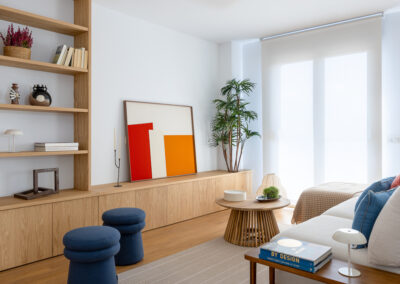 Salón con mueble a medida de madera, estantería abierta, sofá en tonos neutros, pufs azules y decoración con cuadros de colores.