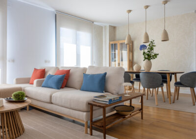 Salón comedor abierto con sofá claro, cojines en azul y terracota, mesa de comedor de madera y sillas tapizadas en tonos neutros.